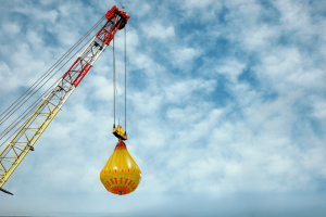 Crane lifting Fleximake water weight bag during load testing, showcasing single-point lift system with yellow PVC bag suspended by master link and webbing straps in an offshore or construction environment.
